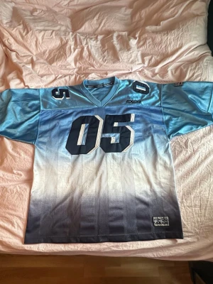 Blå jersey från EXCO - Säljer en oversized mesh jersey topp från EXCO i blått med stora vita och mörkblå siffror '05' fram och bak. Tröjan har v-ringad krage, korta ärmar och cool gradient från ljusblå till mörkblå. Perfekt för sportig streetstyle.