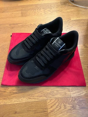 Valentino Garavani svarta sneakers - Säljer ett par svarta Valentino Garavani sneakers med coola camouflagedetaljer. Skorna har snörning, platt sula med nitar på hälen och är gjorda i en mix av mocka, skinn och textil. Perfekta för dig som vill ha en stilren men edgy look.