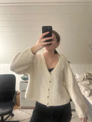 Vit kofta från Gina Tricot i ullmix - Mysig vit kofta från Gina Tricot i ullblandning. Koftan har v-ringning, långa ärmar och knäpps med silvriga knappar framtill. Den är lite croppad och har en loose fit. Super mjuk men har tyvärr fått en del nopprar☹️ funkar ändå att använda såklart och inga fler skavanker än detta🥰