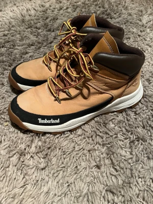 Beige Timberland boots i skinn med snörning - Säljer ett par snygga Timberland boots i beige och svart med bruna detaljer. Skorna har robust snörning med gula och röda skosnören, vadderad brun krage och vit sula. Tillverkade i skinn och mocka, perfekta för höst och vinter.