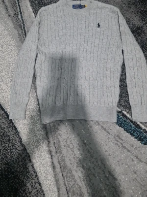 Ljusgrå kabelstickad tröja Polo Ralph Lauren - Säljer en ljusgrå kabelstickad tröja från Polo Ralph Lauren. Tröjan har rund hals, lång ärm och den klassiska broderade loggan på bröstet. Perfekt för dig som gillar stilren och tidlös design.