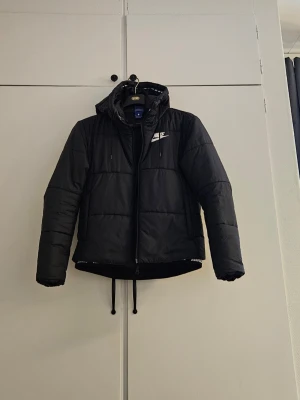 Svart pufferjacka från Nike - Säljer en svart pufferjacka från Nike med huva och vita loggan på bröstet. Jackan har dragkedja framtill och snörning nedtill för extra passform. 2 fickor med dragkedja Perfekt för kalla dagar och riktigt snygg 