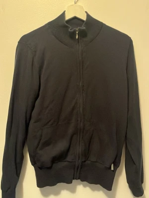 Svart kofta från Alcan med dragkedja - Svart kofta från Alcan med hög krage och hel dragkedja framtill. Tröjan har ribbade muddar vid krage, ärmslut och nederkant samt diskret logga broderad på ryggen. Perfekt för lager på lager under kyliga dagar. Liten i storlek. Skulle säga att den sitter som S