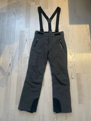 Grå skidbyxor/överdragisar från Swedemount - Skidbyxor/överdragisar i grått från Swedemount med justerbara hängslen som går att ta av och dragkedjor på fickorna. Byxorna har förstärkta partier vid bensluten och kardborre i midjan för bästa passform. Perfekta för kalla dagar och vinteraktiviteter.