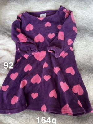 Lila velourklänning med hjärtan - Supermjuk lila velourklänning från Polarn o pyret med rosa hjärtan över hela. Klänningen har långa ärmar med resår vid handlederna och en bekväm passform. Perfekt för mysiga dagar och enkel att ta på tack vare knapp vid axeln.🌸 Fint använt skick. Storlek 92