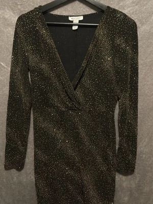 Glittrig svart klänning från NLY One - Supersnygg svart kortklänning från NLY One med massor av guldglittrande detaljer. Djup v-ringning och långa ärmar ger en riktigt festlig vibe. Materialet är stretchigt och sitter snyggt på kroppen. Perfekt för dig som vill sticka ut och glänsa!