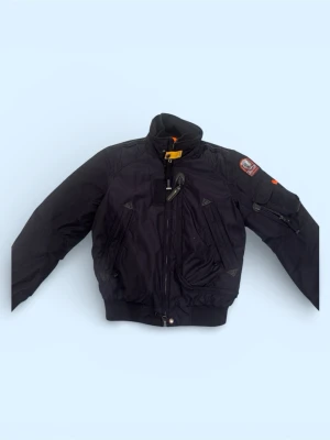 Svart bomberjacka från Parajumpers XS - Säljer en svart bomberjacka från Parajumpers i storlek XS 176cm . Jackan har orange innerfoder, flera dragkedjor, hög krage med gul detalj och patch på ärmen. Tillverkad i slitstark polyester och perfekt för kalla dagar. Snygg och funktionell design. Kvitto finns