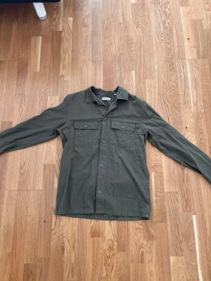 Mörkgrön overshirt från Jack and Jones  - Säljer en stilren mörkgrön overshirt från Zara Man. Skjortjackan har två bröstfickor med knappar, klassisk krage och långa ärmar. Perfekt att bära öppen eller stängd för en avslappnad look.
