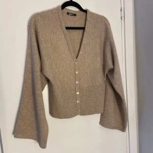 Beige kofta från Gina Tricot - Säljer en beige kofta från Gina Tricot med v-ringning och vida ärmar. Koftan har ribbad nederkant och knäppning med pärlemorknappar framtill. 