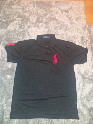 Sportpiké från Polo Ralph Lauren M - Klassisk svart pikétröja från Polo Ralph Lauren med stor röd broderad logga på bröstet och röd detalj på ärmen. Tröjan har krage, knappar framtill och är kortärmad. Tillverkad i polyester