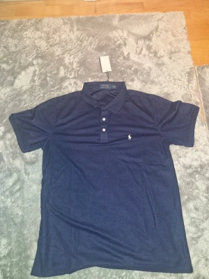 Marinblå pikétröja från Ralph Lauren - Säljer en klassisk marinblå pikétröja från Polo Ralph Lauren. Tröjan har korta ärmar, krage och tre knappar framtill. Den ikoniska vita loggan är broderad på bröstet. Perfekt till jeans eller shorts för en clean och stilren look. Material: Polyester