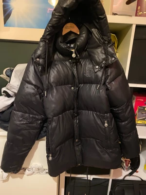 Svart pufferjacka Messi x SikSilk - Svart pufferjacka från Messi x SikSilk med hög krage, huva och dragsko. Jackan har två fickor med dragkedja och broderad logga på bröstet. Perfekt för kalla dagar och riktigt snygg.