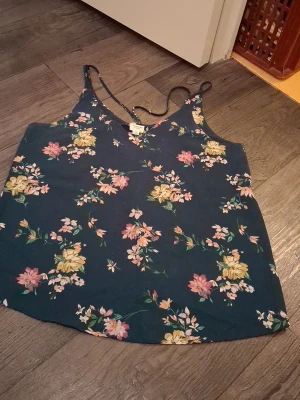 Blommig linnetopp från Jacqueline de Yong - Mörkgrön topp från Jacqueline de Yong med tunna axelband och blommigt mönster i rosa, gult och grönt. Toppen har en lätt v-ringning och är perfekt för varma dagar. Materialet känns lätt och luftigt.