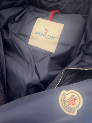 Mörkblå Moncler dunjacka - Säljer en mörkblå dunjacka från Moncler med klassisk logga på ärmen. Jackan har huva, flera dragkedjeförsedda fickor och quiltade detaljer. Tillverkad i polyester och dun, perfekt för kalla dagar. Snygg och tidlös design med en schysst passform.