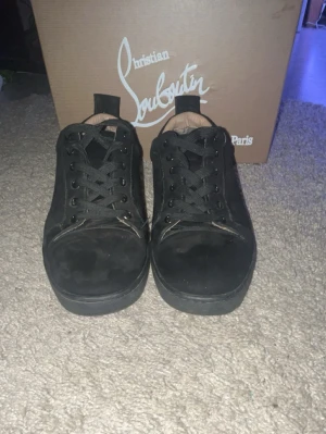 Svarta sneakers från Christian Louboutin - Säljer ett par svarta sneakers från Christian Louboutin med klassisk röd sula. Skorna har snörning, rund tå och är tillverkade i mocka. Insidan är beige och designen är stilren och lyxig, perfekt för dig som vill sticka ut med exklusiva detaljer.