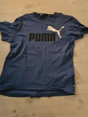 Blå Puma t-shirt med logga - Snygg blå t-shirt från Puma med stor vit logga och svart text på bröstet. Klassisk passform och rund hals. Tillverkad i mjuk bomull som känns skön mot huden. Perfekt för dig som gillar sportig och enkel stil.