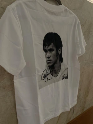 Neymar T-Shirt - Lyft din stil med vår vit premium T-shirt med ett svartvitt tryck av Neymar på ryggen. En stilren och kraftfull design som hyllar en av världens största fotbollsstjärnor – perfekt för både vardag och streetwear-look. T-shirten är tillverkad i mjuk och slitstark bomull som ger hög komfort hela dagen. Den minimalistiska framsidan kombineras med ett iögonfallande motiv på ryggen, vilket skapar en modern och trendig kontrast  