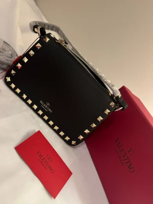 Svart axelväska  - Säljer en svart axelväska från med guldfärgade nitar runt kanterna och en praktisk dragkedja upptill. Väskan är tillverkad i skinn och har en stilren, rektangulär form. Perfekt för dig som gillar exklusiva detaljer och snygg design.