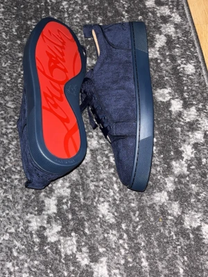 Mörkblå sneakers från Christian Louboutin - Säljer ett par snygga mörkblå sneakers från Christian Louboutin med klassisk röd sula och signatur på undersidan. Skorna har snörning och är tillverkade i ett mjukt tygmaterial med detaljer i gummi. Perfekt för dig som vill sticka ut med exklusiva och stilrena sneakers.
