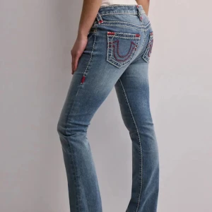 True Religion jeans med kontrastsömmar - Snygga blå jeans från True Religion med ikoniska kontrastsömmar i vitt och rött. Modellen har bootcut-ben, låg midja och klassiska bakfickor med broderad logga. Jeansen är i denim och har detaljer som synliga sömmar och logopatch bak.
