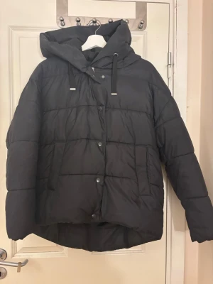 Svart pufferjacka från Vero Moda - Svart pufferjacka från Vero Moda med huva och snörning. Jackan har knappar framtill och två sidofickor. Den är quiltad och har en loose passform, perfekt för kalla dagar. Materialet är polyester och jackan är långärmad. Den har ett litet hål på framsidan men inget man tänker på.