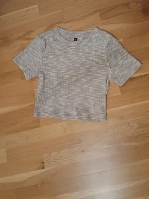 Ribbad beige croppad topp H&M Divided - Kortärmad croppad topp från H&M Divided i beige nyans med ljusa inslag. Ribbad struktur och rund halsringning ger en enkel men trendig look. Perfekt till jeans eller kjol för en avslappnad stil.