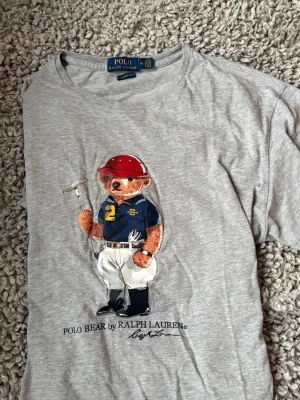 Grå Polo Bear t-shirt från Ralph Lauren - Säljer en grå t-shirt från Polo Ralph Lauren med den ikoniska Polo Bear-trycket framtill. T-shirten har rund hals och korta ärmar. Materialet är mjuk bomull och passformen är klassisk. Perfekt för dig som gillar streetwear och vill ha något med lite extra attityd.