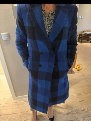 Blårutig lång kappa från Lindex XS - Säljer en snygg lång kappa från Lindex i storlek XS. Den har ett blårutigt mönster med svarta inslag, dubbelknäppning framtill och klassisk krage. Materialet är ullblandning som känns mjukt och värmande. 