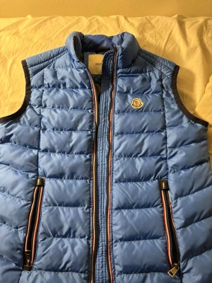 Blå dunväst från Moncler - Snygg blå dunväst från Moncler med quiltad design och svarta detaljer. Västen har dragkedja framtill, två fickor med dragkedja och Moncler-logga på bröstet. Utsidan är blå och insidan svart, vilket ger en clean kontrast. Perfekt för lager-på-lager-stil.