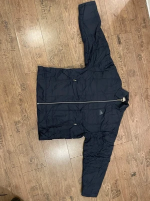 Svart pufferjacka från Adidas - Säljer en svart pufferjacka från Adidas med diskret rutmönster och dragkedja framtill. Jackan har två fickor med dragkedja och hög krage. Jackan är knappt använd och har inga skador på sig