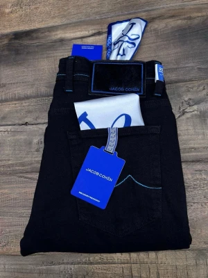 Svarta jeans från Jacob Cohën - Snygga svarta jeans från Jacob Cohën med blå kontrastsömmar och exklusiva detaljer. Jeansen har klassisk femficksdesign, blå etikett och medföljande scarf. Materialet är mjukt och stretchigt jeans, perfekt för en modern look.