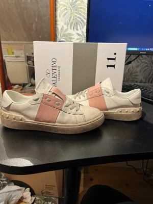Valentino Garavani Open sneakers rosa/vit - Säljer ett par Valentino Garavani Open sneakers i vit och ljusrosa skinn med coola nitar på hälen och platt sula. Skorna har snörning och en bred rosa panel över mitten. Märkeslogga på plösen och box medföljer. Tvättat skorna innan jag skickar iväg dom!