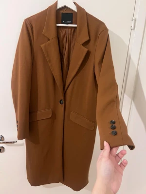 Brun kappa från Amisu med knappar - Säljer en snygg brun kappa från Amisu med klassisk krage och svarta knappar både framtill och vid ärmsluten. Kappan har två fickor med lock och en rak, stilren passform som passar perfekt till lager-på-lager. Perfekt för dig som gillar en clean och tidlös look.Passar en M -XL i storlek