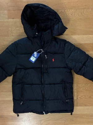 Svart pufferjacka från Polo Ralph Lauren - Säljer en svart pufferjacka från Polo Ralph Lauren med huva och dragkedja. Jackan har det klassiska röda Polo-logot på bröstet, två sidofickor med dragkedja och en bröstficka. Perfekt för kalla dagar och riktigt snygg till jeans.