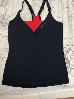 Svart och röd Better Bodies linnetopp - Säljer en svart linnetopp från Better Bodies med röd kontrastdetalj vid bröstet och justerbara axelband. Toppen har en sportig look och är tillverkad i ett stretchigt syntetmaterial som passar perfekt till träning. Loggan sitter diskret på bröstet.