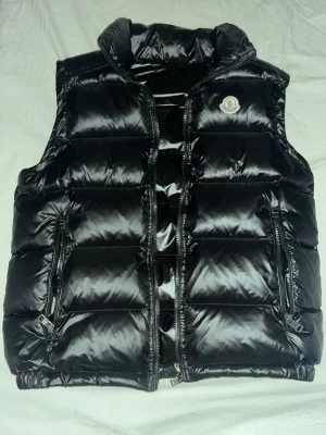 Svart dunväst från Moncler - Säljer en svart, glansig dunväst från Moncler med hög krage och två dragkedjeförsedda fickor framtill. Västen har quiltad design och klassisk Moncler-logga på bröstet. Fick den av min bror som flytta fläckfri och nfc koden scannar 