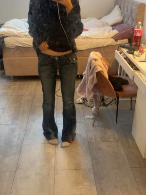 True Religion blå bootcut jeans - Säljer ett par ikoniska True Religion jeans i mörkblå tvätt med kontrastsömmar och snygga detaljer på bakfickorna. Modellen är lågmidjad med bootcut och klassiska knappar. Jeansen har slitningar och är tillverkade i stretchig bomull.