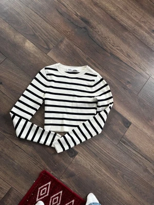 Randig croppad topp från Zara - Säljer en vit och svart randig croppad topp,magtröja. från Zara. Toppen har lång ärm och rund halsringning, med ett stilrent och klassiskt mönster. 