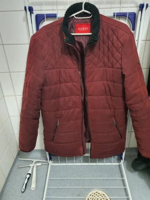 Vinröd pufferjacka Guess XL - Säljer en vinröd pufferjacka från Guess i storlek XL. Jackan har svart teddyfoder i kragen, dragkedja framtill och två fickor med dragkedja. Materialet är polyester och jackan har en schysst, modern look som passar perfekt till höst och vinter. Kedjan behövs bytas ut, den fungerar ej