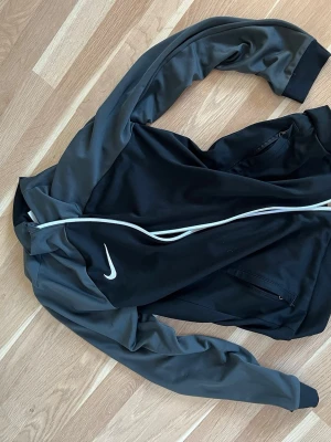 Nike svart träningsjacka slim fit - Nike träningsjacka i svart med grå ärmar och vit dragkedja. Jackan har Dri-Fit material som andas och en snygg slim fit passform. Klassisk Nike-logga på bröstet och praktiska fickor med dragkedja. Perfekt för träning eller chill.