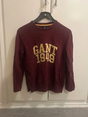 Vinröd stickad tröja från GANT - Säljer en vinröd stickad tröja från GANT med broderad gul text 'GANT 1949' på bröstet. Tröjan har rund halsringning, ribbade muddar och lång ärm. Perfekt för dig som gillar klassisk college-stil.
