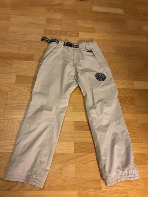 Beiga skidbyxor från Dope Snow - Säljer ett par beiga skidbyxor från Dope Snow med svart logga på benet och justerbart midjeband med tryck. Byxorna har elastiska benslut med knappar och praktiska fickor. Perfekta för skidsäsongen.