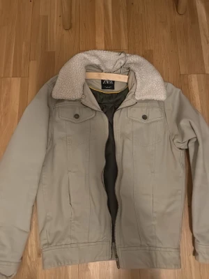 Beige jacka från Zara - Går ner i pris vid snabba affär. Säljer en beige teddyjacka från Zara med mjuk, vit fuskpäls på kragen och dragkedja framtill. Jackan har två bröstfickor med knappar och en klassisk, rak passform. Perfekt för dig som vill ha en stilren och mysig jacka till hösten eller vintern. 