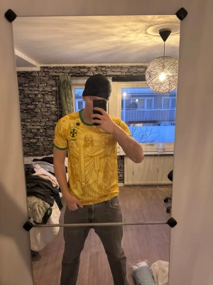 Gul Brasil Nike t-shirt med tryck - Säljer en gul Brasil t-shirt från Nike med gröna detaljer och stort grafiskt tryck av Kristusstatyn och brasilianska motiv. Klassisk rund halsringning och korta ärmar. Perfekt för dig som vill sticka ut med en unik och färgstark tröja.