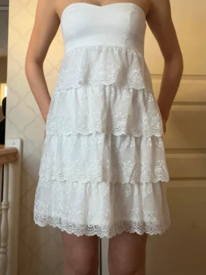 Vit axelbandslös spetsklänning - Superfin vit kortklänning med flera lager volanger och spetsdetaljer. Axelbandslös modell med söt hjärtformad överdel och broderade blommor på kjolen. Perfekt för sommarens alla tillfällen!
