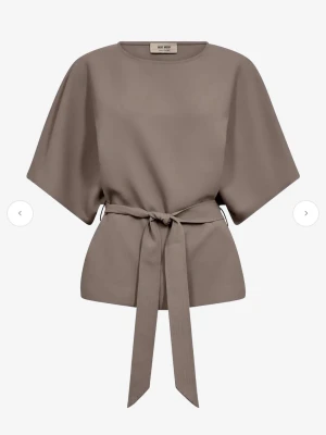 Beige blus med knyt från Mos Mosh - Säljer en stilren beige blus från Mos Mosh med breda korta ärmar och ett snyggt knytband i midjan. Blusen har rund halsringning och en lös passform som ger en avslappnad men chic look. Perfekt till jeans eller kjol för en trendig outfit. Helt ny och oanvänd