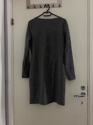 Glittrig svart kortklänning - Säljer en svart glittrig kortklänning med lång ärm och djup rundringning på ryggen . Klänningen har en enkel, rak passform och är perfekt för dig som vill sticka ut. Materialet är stretchigt och skimrar snyggt i ljuset.