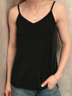 Glittrig svart linnetopp Vero Moda - Säljer en svart glittrig linnetopp från Vero Moda med tunna axelband och v-ringad hals. Toppen har en lätt skimrande yta och är perfekt för dig som gillar att sticka ut lite extra. Materialet känns mjukt och följsamt mot huden.