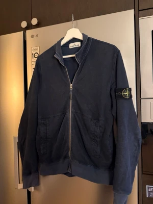 Marinblå ziptröja från Stone Island - Säljer en marinblå ziptröja från Stone Island med ikonisk patch på ärmen. Tröjan har hel dragkedja framtill, två fickor och ribbade muddar. Tillverkad i mjuk bomull och passar perfekt till lager på lager under höst och vår. Såklart äkta!!