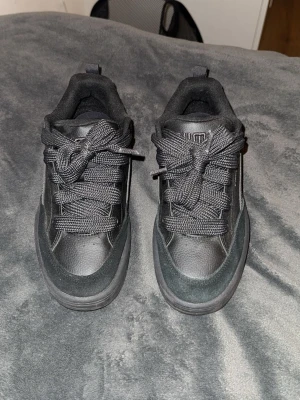 Svarta Puma sneakers i skinn och mocka - Säljer ett par svarta Puma sneakers med chunky sula och breda snören. Skorna är gjorda i en mix av skinn och mocka, med klassisk Puma-logga på sidan. Låg modell med rund tå och platt sula, perfekta till streetwear eller casual outfits.